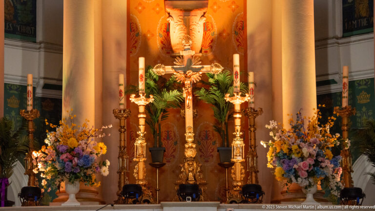 Crucifix surrounded by tall gilded candlesticks, flowers, greenery, and columns in the Basilica-Cathedral sanctuary.