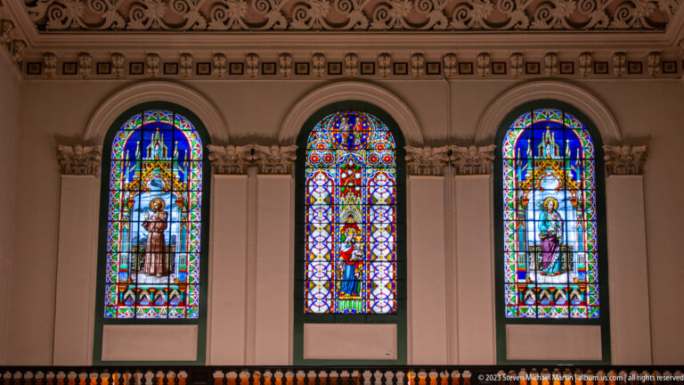 Three tall arched stained-glass windows with sacred figures, colorful borders, carved capitals, and decorative molding.