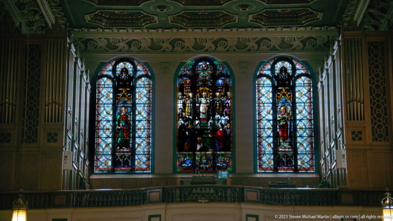 Three stained-glass windows above a dark interior balcony, flanked by tall woodwork beneath a patterned ceiling.
