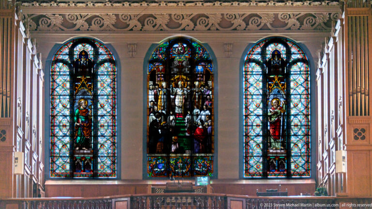 Close view of three colorful figural stained-glass windows above a wooden balcony rail in the Basilica-Cathedral.