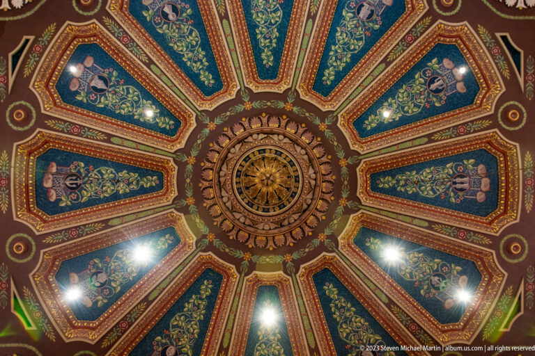 Centered view of an ornate radial cathedral ceiling with circular boss and alternating blue-green and red painted panels.