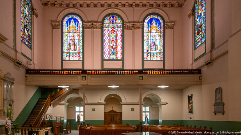 Three stained-glass windows above a gallery, with wooden stair, green lower walls, arches, and pews below.
