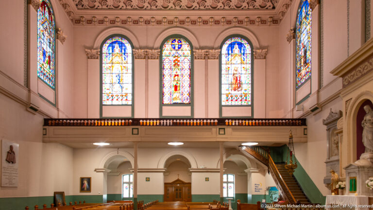 Rear cathedral gallery with three stained-glass windows, central doorway, arches, stair, and a side devotional niche.
