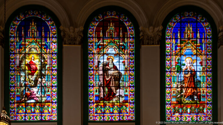 Three brightly colored stained-glass windows with sacred figures beneath Gothic-style canopies and patterned borders.