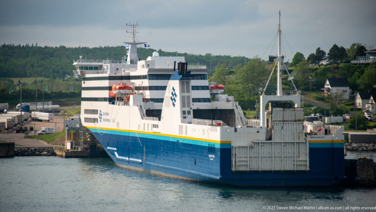 Blue-and-white Marine Atlantic ferry viewed from the water at a terminal, with its stern loading structure facing the harbor.