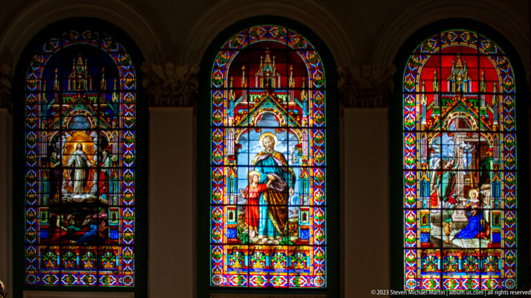 Two colorful arched stained-glass windows showing sacred figures and narrative scenes beneath elaborate canopies.