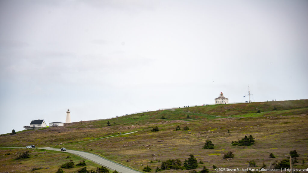 Road winding across low coastal vegetation toward lighthouse structures on the open Cape Spear headland.