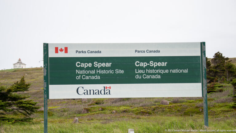 Bilingual Parks Canada sign for Cape Spear National Historic Site with the historic lighthouse visible on the hill behind.