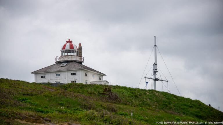 Historic Cape Spear lighthouse with white keeper’s residence, hipped roof, and red-and-white lantern on a grassy hill.