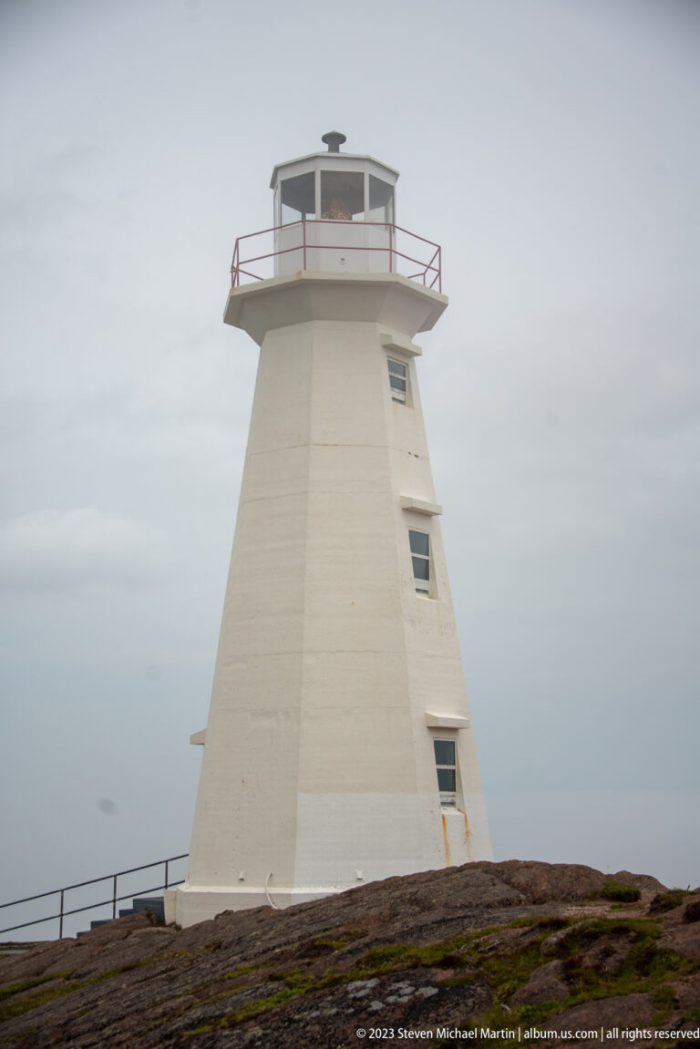 Low-angle view of a tapered white Cape Spear lighthouse tower with polygonal lantern room and red gallery railing.