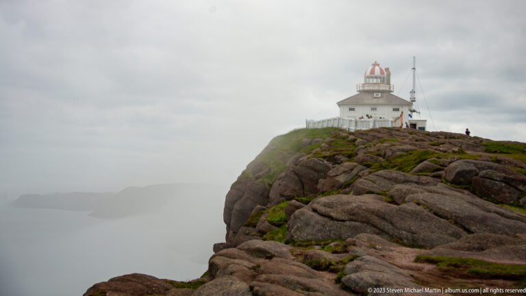 Historic Cape Spear lighthouse above broad fractured bedrock, with fog obscuring the ocean and distant coast.