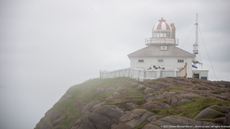 Historic Cape Spear lighthouse in fog, with white clapboard residence, red-and-white lantern, fence, and nearby mast.