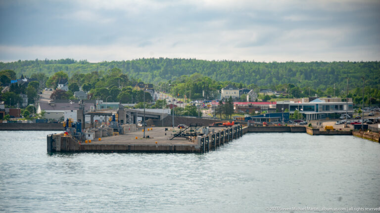 Concrete ferry terminal pier with loading equipment and low waterfront buildings, viewed across calm harbor water.