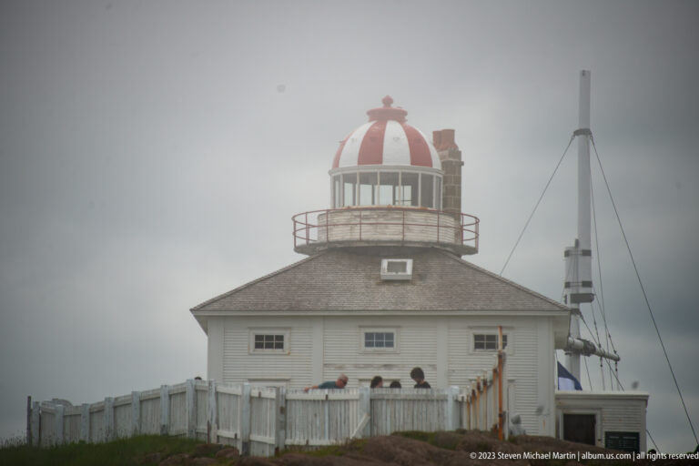 Front view of historic Cape Spear lighthouse with striped lantern, white keeper’s residence, fence, visitors, and mast.