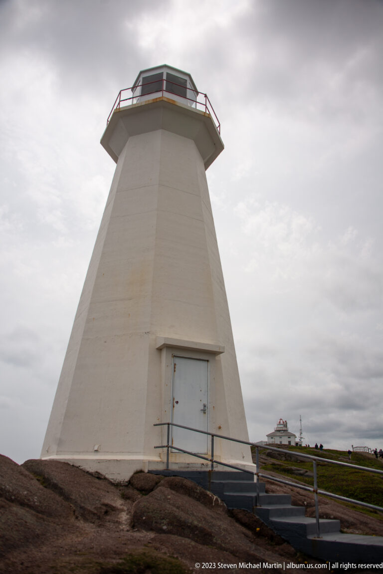 White Cape Spear lighthouse tower with entrance door and access ramp; historic lighthouse appears on the distant ridge.
