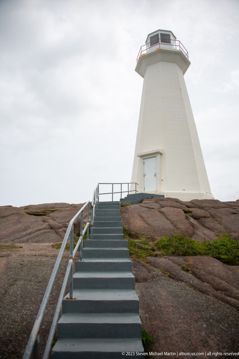 Gray stairs lead directly across exposed bedrock to the entrance of the tapered white Cape Spear lighthouse tower.