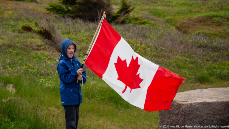 Hooded visitor holding a large windblown Canadian flag beside low coastal vegetation at Cape Spear.