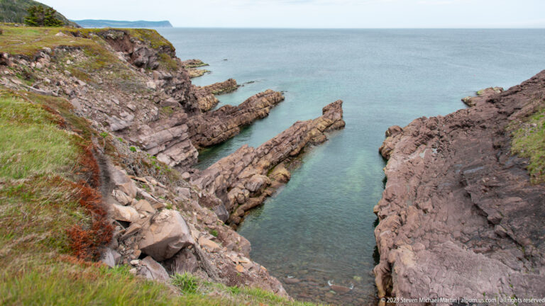 Narrow turquoise inlet between fractured reddish-gray rock walls opening toward the Atlantic at Cape Spear.