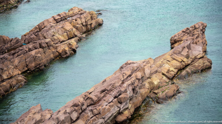 Parallel reddish-brown bedrock ribs project into clear blue-green seawater along the rocky coast near Cape Spear.
