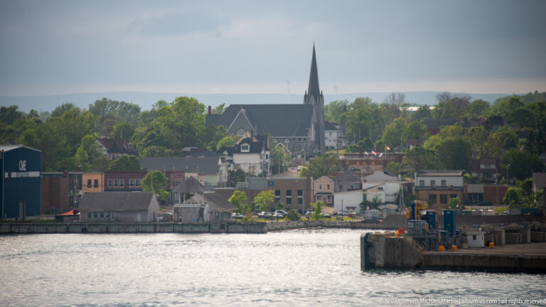 Dark steep-roofed church with a tall pointed steeple rises above waterfront buildings across a harbor.
