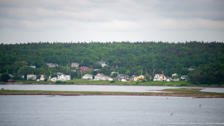 Small houses line a distant shore below a forested ridge, with shallow water and exposed tidal ground in front.
