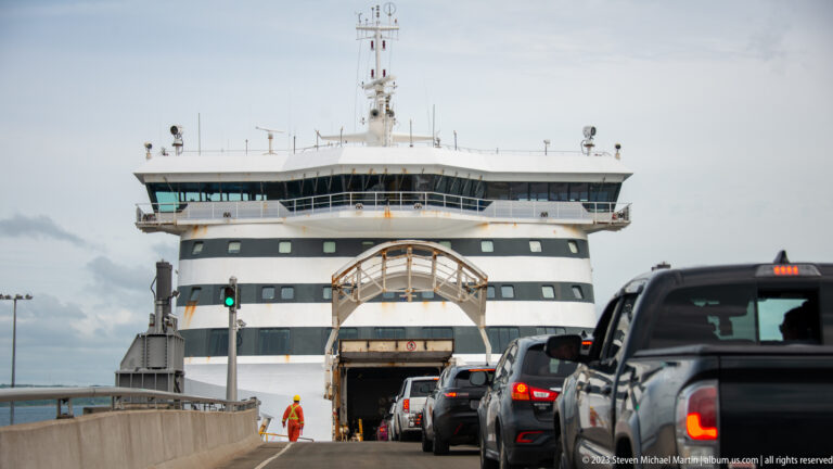 Cars line up at the open stern loading door of a large ferry while a crew member stands beside the lane.