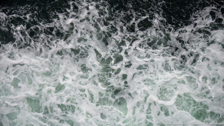 Dense white foam and swirling green-gray seawater fill the frame in the ferry's turbulent wake.