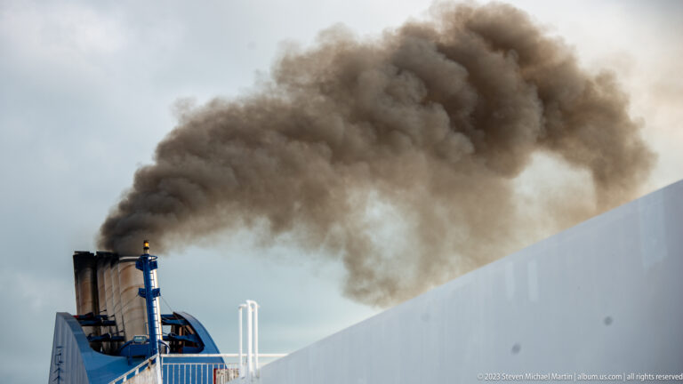 Thick dark exhaust smoke streams from a ferry funnel above the white ship superstructure.