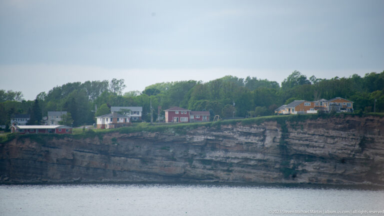 Several houses stand above a horizontally layered rocky sea cliff beside calm water.