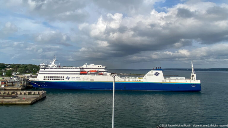 Large blue-and-white Marine Atlantic ferry lies broadside at a harbor berth beneath dramatic clouds.
