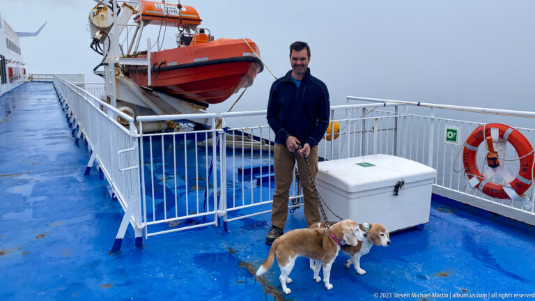 Man holds two beagles on a wet blue ferry deck beside an orange rescue craft and life ring in fog.
