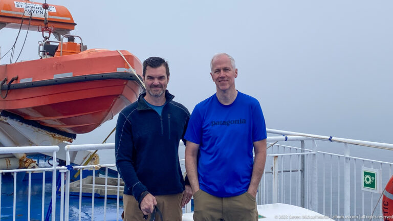 Two men pose on a ferry deck beside an orange rescue craft and white railings in misty weather.