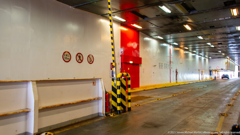 Long empty vehicle deck inside a ferry with steel walls, overhead lights, lane markings, and striped protective posts.