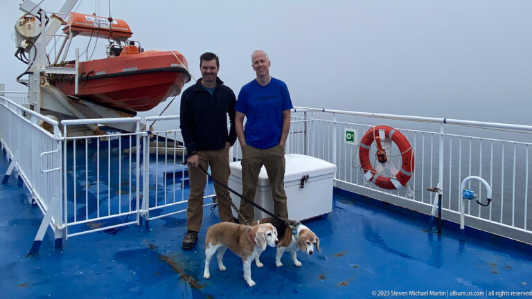 Two men stand with two beagles on a wet ferry deck between an orange rescue craft and a life ring in fog.