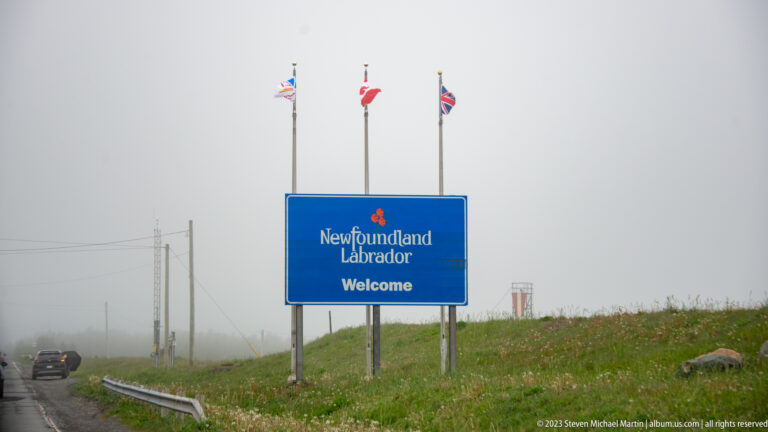 Blue Newfoundland and Labrador welcome sign beside a foggy highway, with three flags flying above the roadside marker.