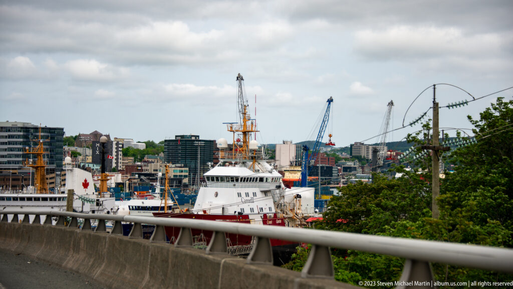 White-and-red working vessel below a roadway, with cranes and waterfront buildings in St. John’s harbor.