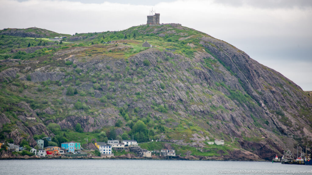 Steep rocky hillside above St. John’s harbor with shoreline houses and a structure on the summit.