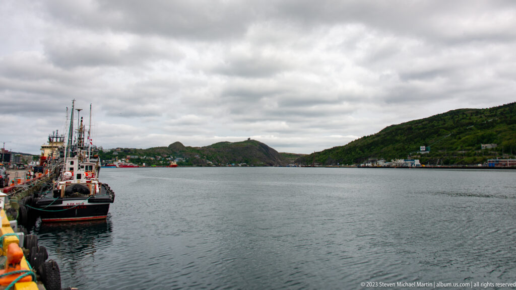 Working boats line a quay beside the open water and steep hills of St. John’s harbor.