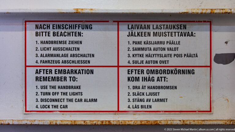 Weathered red-bordered ferry sign with after-embarkation vehicle instructions printed in German, Finnish, English, and Swedish.