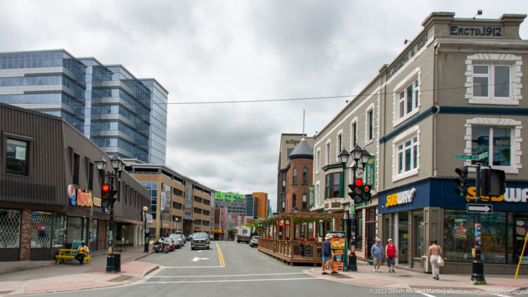 Downtown St. John’s street with shops, pedestrians, traffic lights, patio seating, and mixed old and modern buildings.