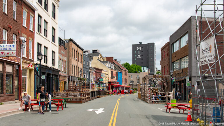 Narrow downtown street lined with colorful pubs, restaurant patios, outdoor tables, and pedestrians in St. John’s.