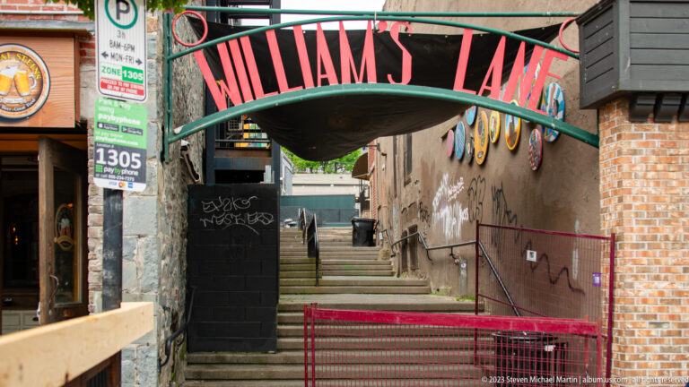 Concrete steps rise through a narrow passage beneath a curved William’s Lane sign between downtown buildings.