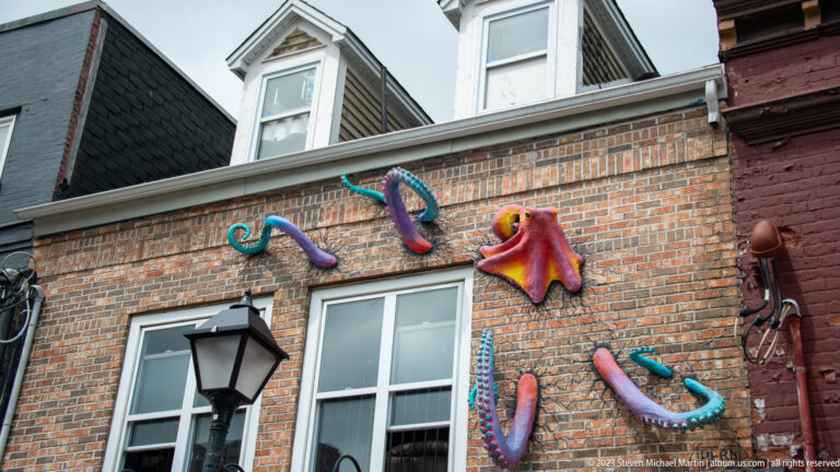Colorful sculpted octopuses with curling tentacles mounted on a brick wall above a window and streetlamp.
