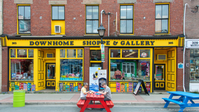 Bright yellow Downhome Shoppe & Gallery storefront at 303–305 with colorful panels and picnic tables outside.