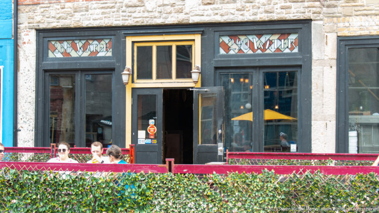 Stone restaurant façade with dark doorway, gold trim, stained-glass transoms, and diners behind a red patio railing.