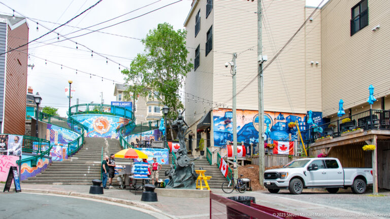 Wide outdoor stairs climb between buildings amid colorful murals, railings, flags, patios, and street activity.