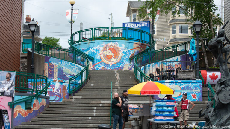 Head-on view of broad downtown stairs with colorful murals, green railings, pedestrians, and a vendor umbrella.
