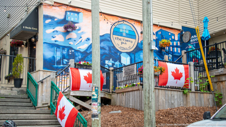 Raised patio at The Republic Pub with a blue-and-orange mural, Canadian flags, stairs, flowers, and blue umbrellas.