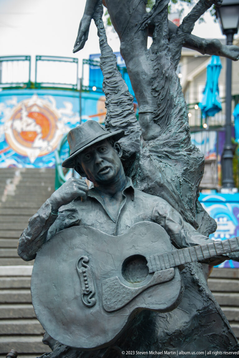 Bronze seated musician in a flat cap holds an acoustic guitar beside a tree-like sculptural form.