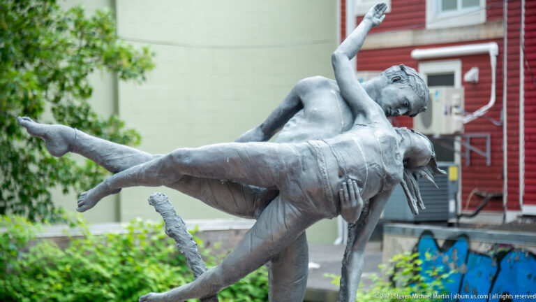 Silver-gray sculpture of two human figures in an acrobatic pose, one held horizontally above the other.